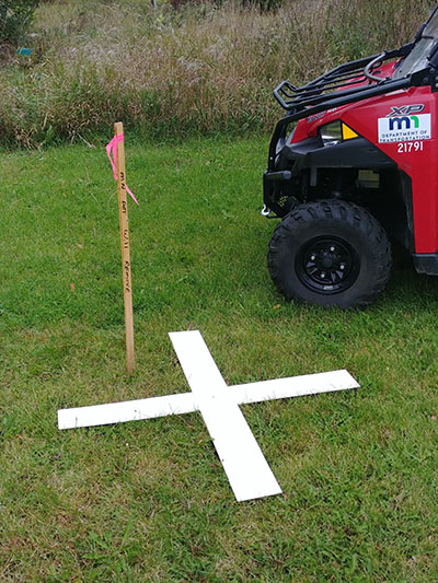 an aerial photography target, consisting of a white X on the ground and a vertical stake with information written on it. An ATV is parked next to it.